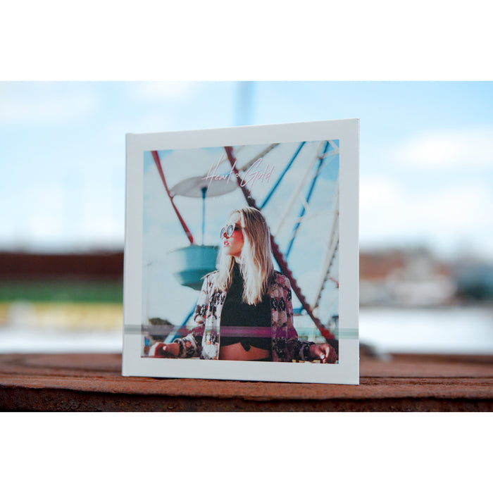 Image of a girl wearing sunglasses and standing in front of a ferris wheel. There is text above the girl that says HEART OF GOLD in pink font, and below that there is text that says BLAME. It is standing on top of a wooden surface.