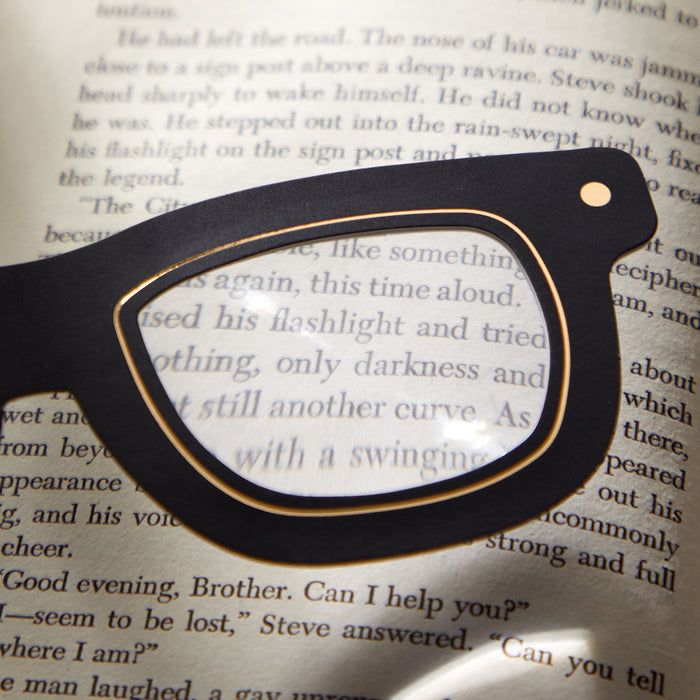 The Galison Classic Readers Bookmark with Magnifier, shaped like black eyeglasses, sits on an open book and stylishly magnifies and highlights text through one lens.