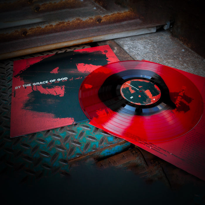 Red vinyl jacket with white text that says BY THE GRACE OF GOD. Behind the text is a black dollar sign covering the whole jacket. On the right side of the jacket is a red and black vinyl. Both are laying on top of an industrial surface.