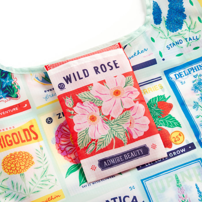 The Emily Taylor Grow Your Own Way Packable Nylon Tote Bag is a colorful, eco-friendly bag featuring a vibrant floral seed packet design with a pink Wild Rose labeled Admire Beauty and illustrations of other flower seed packets.
