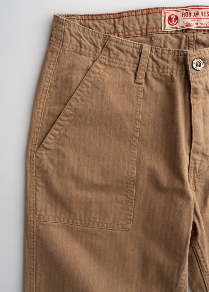Close-up of brown pants with a front pocket, button, and visible stitching. A label reads "Iron and Resin Premium Goods."