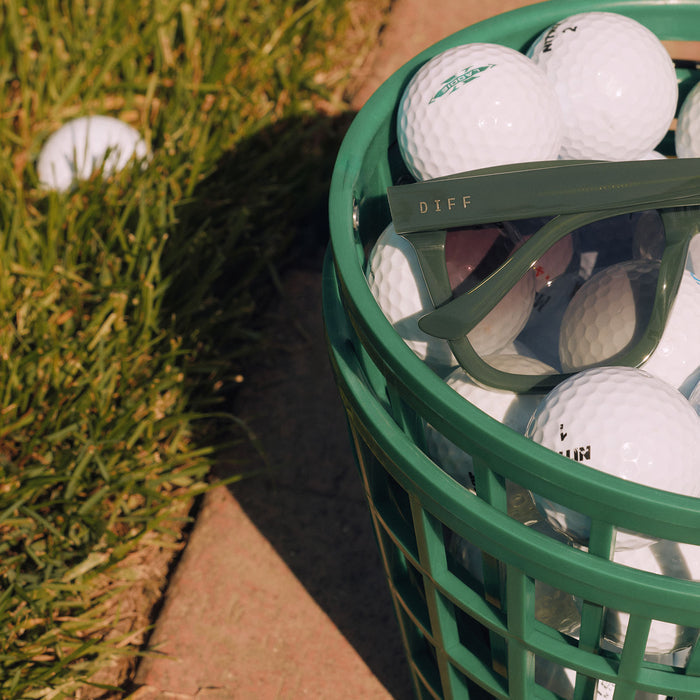 still life - diff eyewear meredith ii square oversized sunglasses with a fairway green acetate frame and grey gradient lenses resting in a golf ball basket