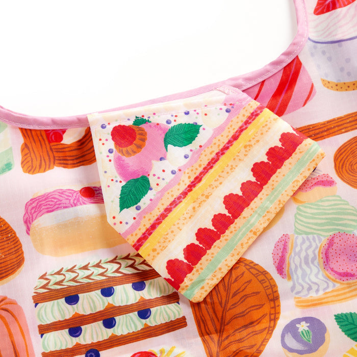A close-up of the Emily Taylor Sweet Confections Packable Nylon Tote Bag, featuring colorful cakes, macarons, and pastries on a light background with a pink neckline trim.