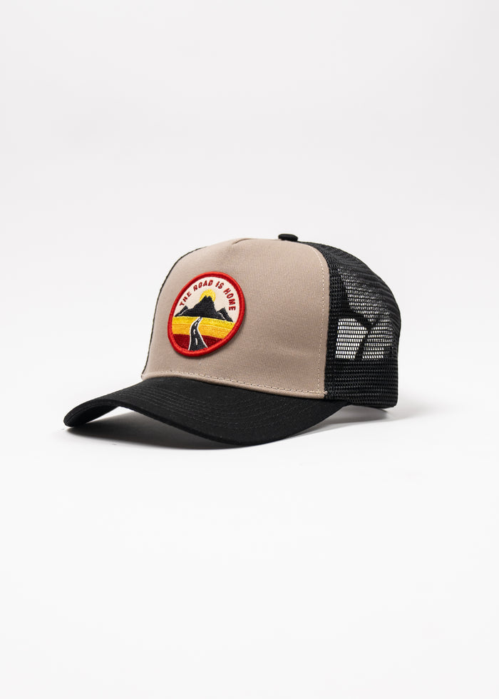 A beige and black trucker hat with a patch reading "The Road is Home" featuring a mountain and road design on the front.