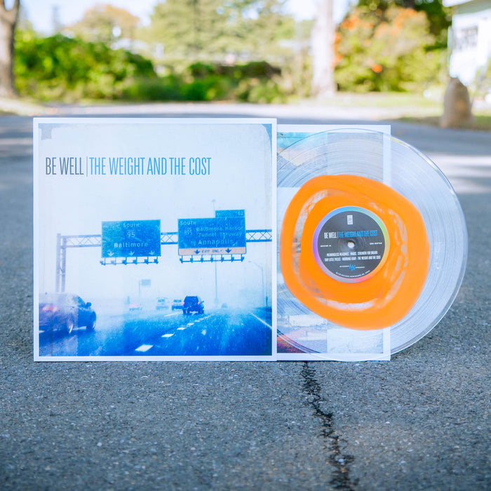 Clear vinyl with thick orange ring towards the center. Vinyl jacket has picture of highway with cars. "BE WELL" is printed in black and "THE WEIGHT AND THE COST" is printed in blue towards top of the jacket.