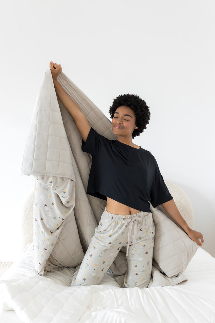 Person holding a beige Harry potter patterned Adult blanket on a bed wearing beige pajamas in harry potter print and black shirt