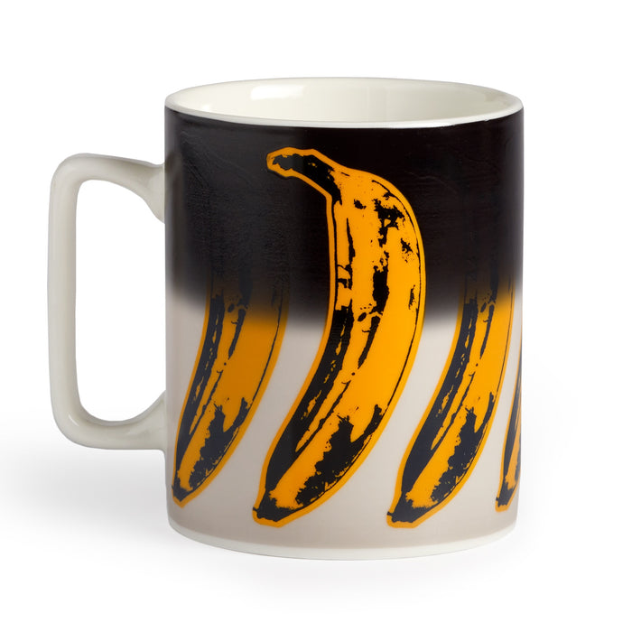 The Andy Warhol Banana Color Changing Ceramic Mug by Andy Warhol features a wraparound pop art design of large yellow bananas with black outlines on a gradient black-to-white background.