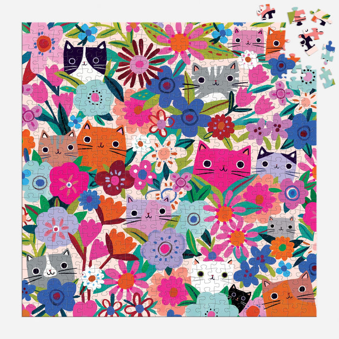 Tara Lilly’s Blooming Cats 500 Piece Puzzle showcases playful cartoon cat faces nestled among vibrant flowers and leaves. A few pieces are missing from the top right corner.