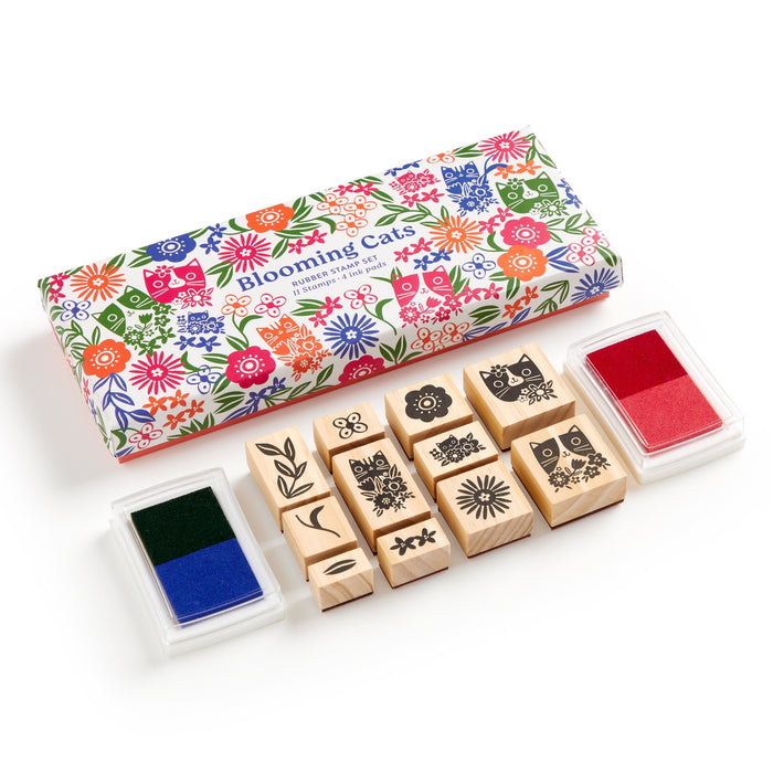 A Tara Lilly Blooming Cats Rubber Stamp Set, featuring nine wooden stamps with botanical and cat designs plus red and blue-green ink pads, comes in a colorful box. The ink pads sit on either side of the vibrant packaging.