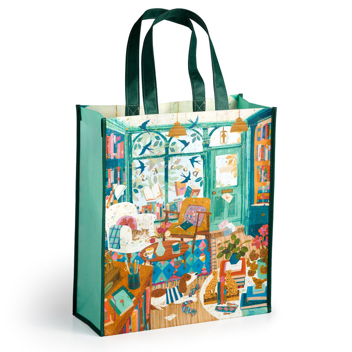 The Bookshop Afternoon Reusable Shopping Bag by Victoria Ball features illustrated art of a cozy studio—bookshelves, plants, art supplies, and a cat—with dark green handles for eco-friendly style.