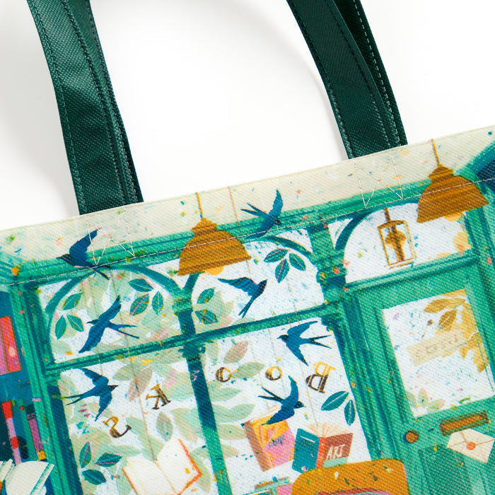 The Bookshop Afternoon Reusable Shopping Bag by Victoria Ball features a vibrant, illustrated scene with blue birds, hanging lights, leafy patterns, and windows. This sustainable tote has green handles and a whimsical, colorful design.