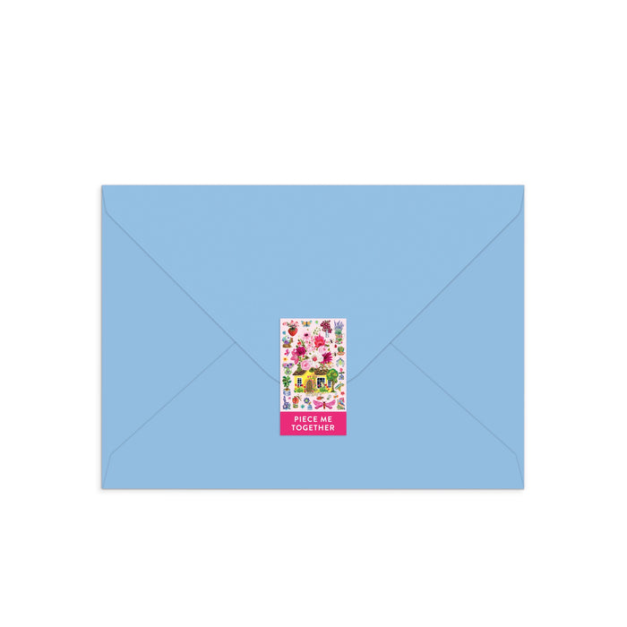 A blue envelope sealed with a vibrant rectangular sticker of floral designs, a yellow house, and “PIECE ME TOGETHER” on pink—a charming Buzzy Bouquets Greeting Card Puzzle by Jennifer O Lewis/August Wren.