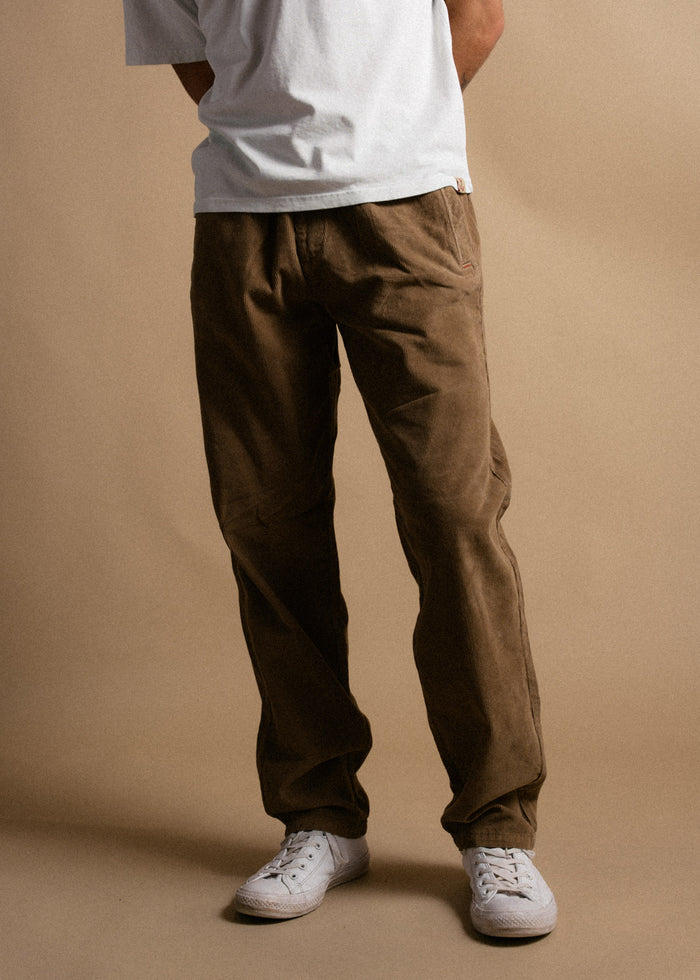 Person in a white shirt and brown pants stands against a beige background, wearing white sneakers. Hands are in pockets.