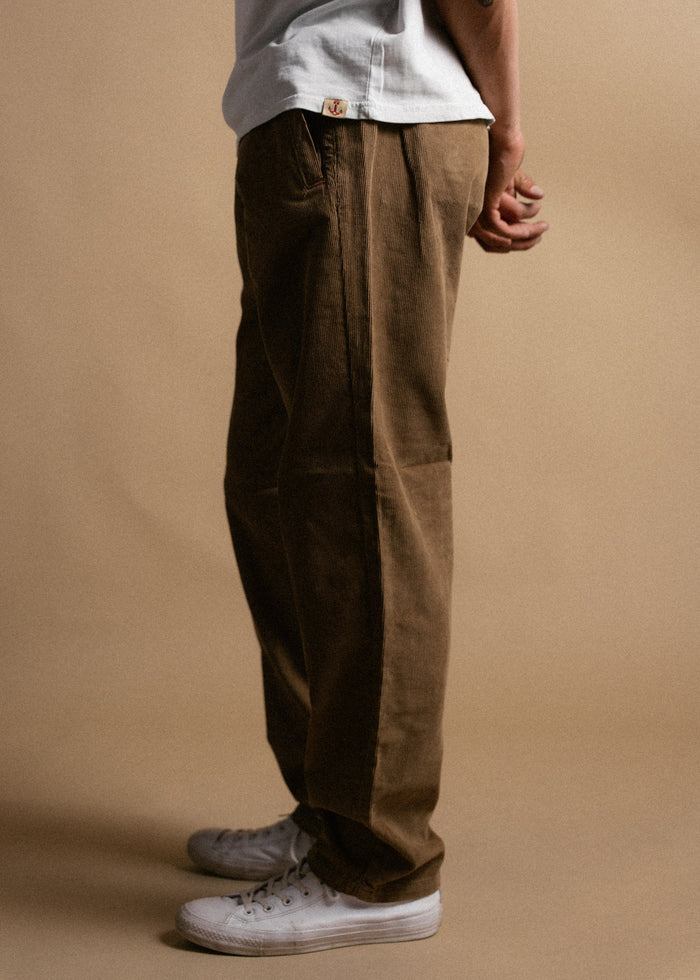Person in brown pants and white sneakers stands against a beige background, wearing a light shirt with a visible tag.