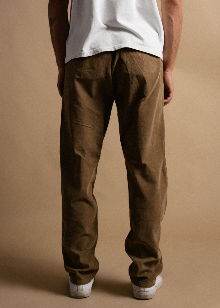 Man in brown corduroy pants and white sneakers stands on a beige background, wearing a white t-shirt.