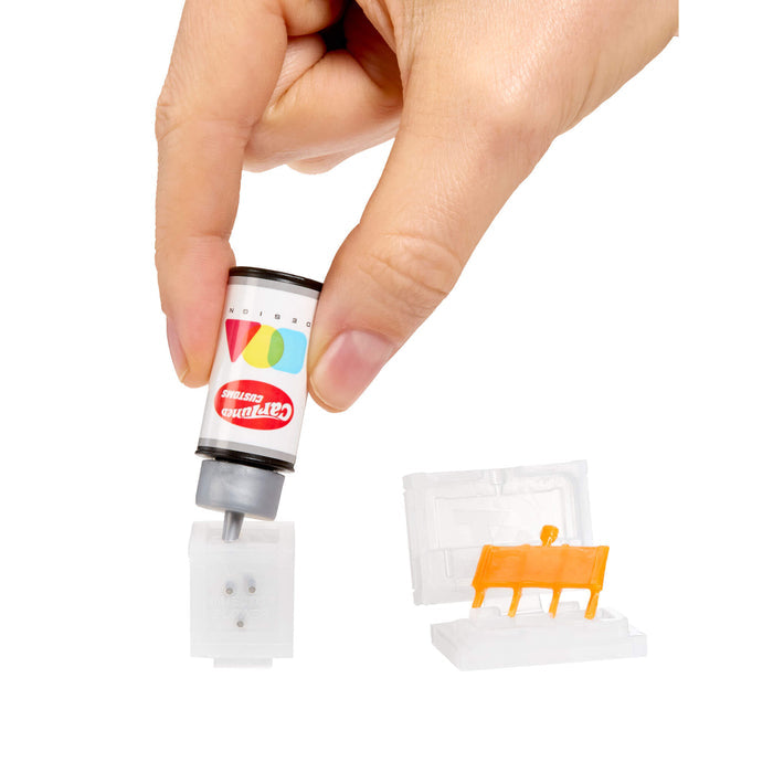 Hand holding a small cartridge with colorful label, next to a white block with an orange design on a white background.