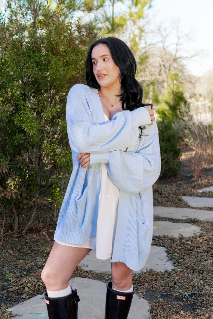 Female model standing outside on a stone path wearing a light cream colored dress with the Chunky Knit Women's Oversized Cardigan in Breeze overtop