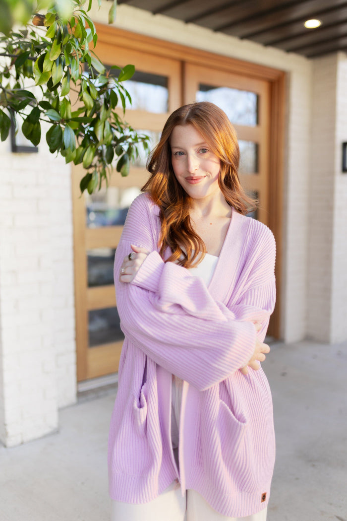 Woman wearing a light purple cardigan standing in front of a wooden door.