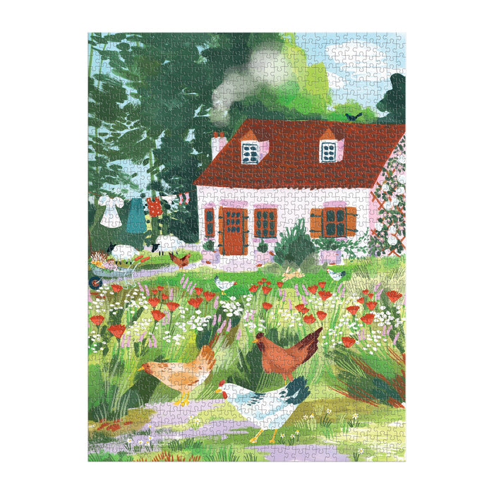 The Joanna Clay Cottage Wildflowers 1000 Piece Puzzle in a Square Box reveals a cozy rural scene with a red-roofed cottage, trees, chickens, laundry on the line, and smoke drifting from the chimney.