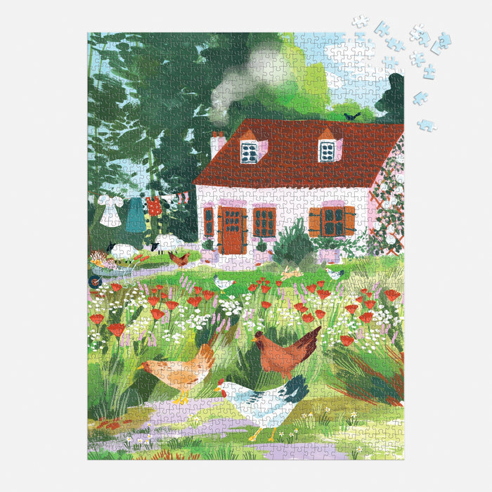 The Cottage Wildflowers 1000 Piece Puzzle in a Square Box by Joanna Clay depicts a charming rural scene with a cottage, chimney smoke, chickens, trees, white laundry, and wildflowers. Some puzzle pieces are missing from the upper right corner.
