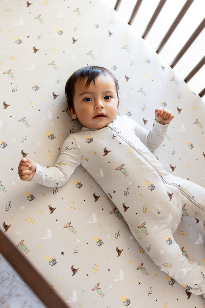 Baby lying on a beige crib sheet with Harry Potter prints, wearing a similar Harry Potter print zippered footie