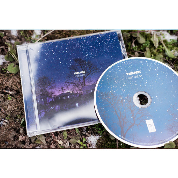 Square CD with an image of a neighborhood at night time. There are stars in the sky and a landscape of houses in the bottom half. In the center, BANE is written in small white letters, and below it is text that says DONT WAIT UP. To the right of the cover is a disc with a similar design, just with trees instead of houses. Both are laying on top of dirt and weeds on the ground.
