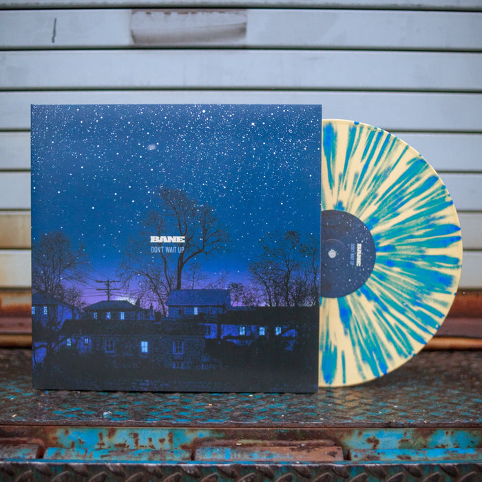 Tan-colored vinyl with blue splatter printed around it. Jacket of vinyl is a picture of a blue and purple night sky with houses, trees, telephone pole, and stars. Center of jacket has "BANE" printed with "DON'T WAIT UP" printed below it. Jacket and vinyl is propped up against a light gray house panel.