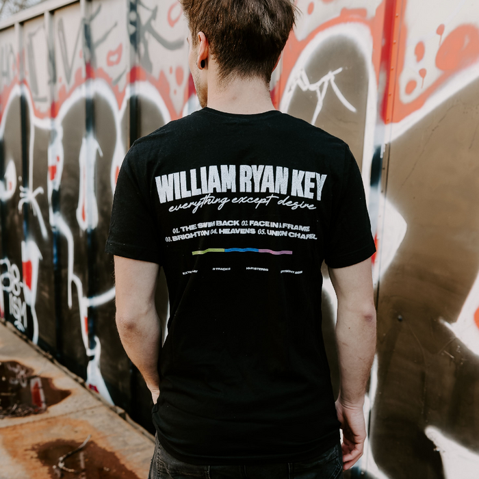 Someone wearing a black short sleeve shirt. There is large white text that says WILLIAM RYAN KEY. Underneath that, there is cursive lettering that says EVERYTHING EXCEPT DESIRE. Below that is a numbered track list. Below that, there is a color blocked line, of green, dark blue, and purple.