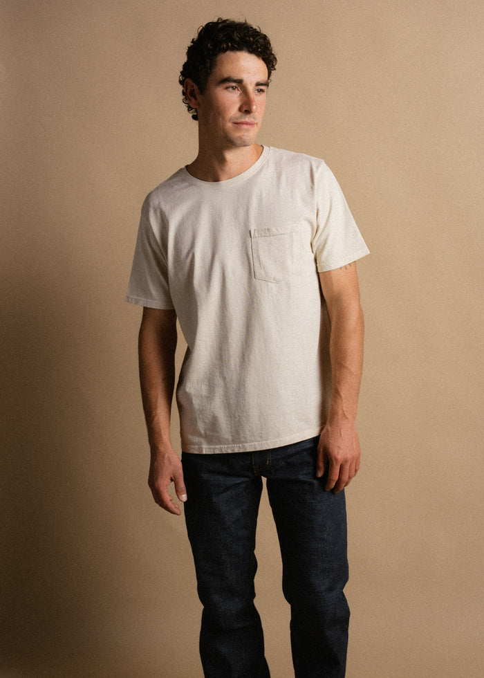 Man in a white t-shirt and jeans stands against a beige background, looking to the side with a neutral expression.