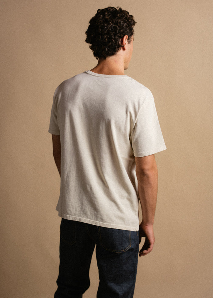 Man in a white t-shirt and jeans stands with his back to the camera against a beige background, showcasing casual fashion.