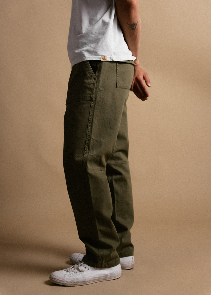 Man in olive green pants, white sneakers, and a white t-shirt stands against a beige background, showcasing casual style.