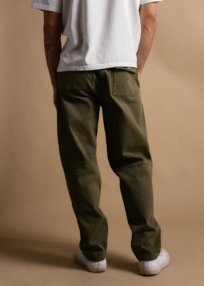 Man in olive pants and white sneakers stands against a beige background, wearing a white shirt, facing away.
