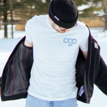 White short sleeve shirt with blue text that says AGO in the top right corner. An individual is modeling the shirt and standing in front of trees. On the ground there is snow.