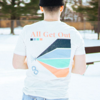 Back of short sleeve shirt with orange text that says ALL GET OUT. Below that is a color blocked design with black, baby blue, teal, and orange coloring. To the left of that design is the same text that says AGO in blue. An individual is modeling the shirt and standing in front of trees. There is snow on the ground.