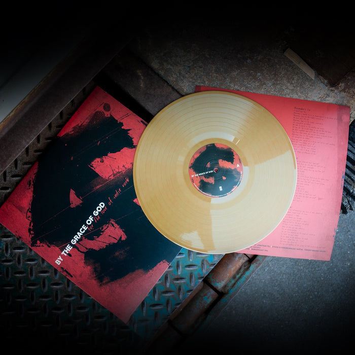 Red vinyl jacket with white text that says BY THE GRACE OF GOD. Behind the text is a black dollar sign covering the whole jacket. On the right side of the jacket is a gold vinyl. Both are laying on top of an industrial surface.