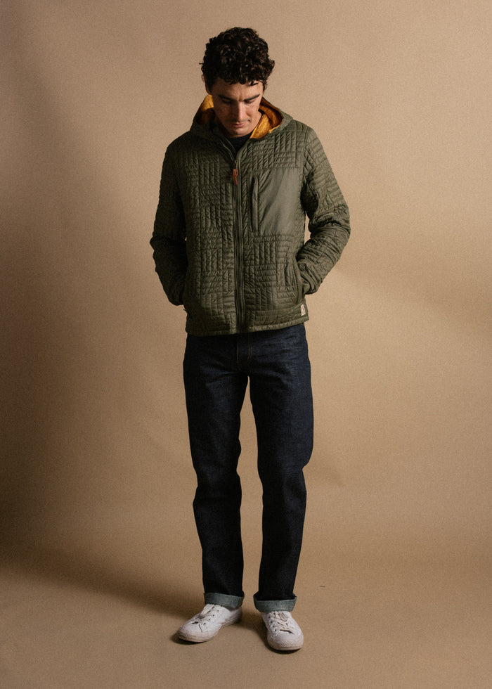 Man in green quilted jacket, dark jeans, and white sneakers stands against a beige backdrop, hands in pockets, looking down.