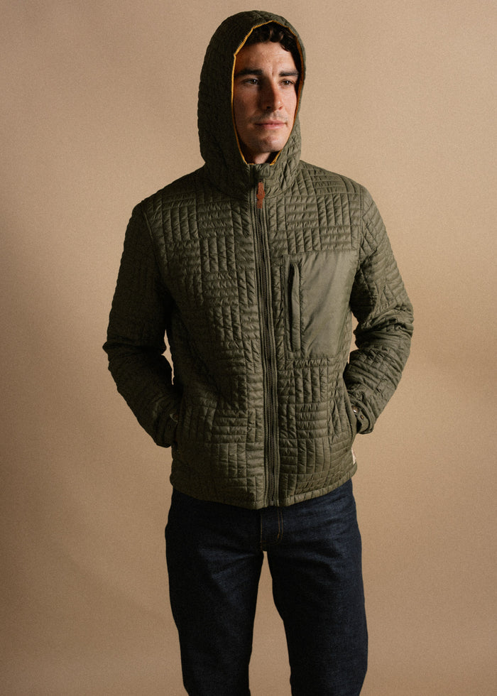 Man in olive quilted jacket with hood up, hands in pockets, stands against a beige background, looking to the side.