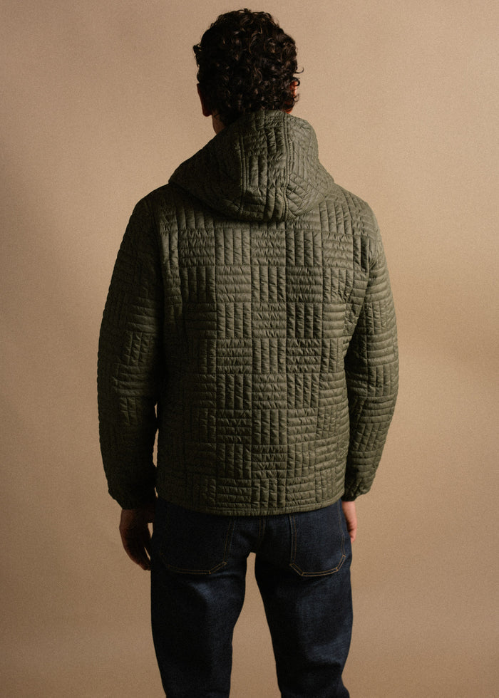 Man in green quilted jacket and jeans stands facing away, against a beige background, highlighting the jacket's texture.