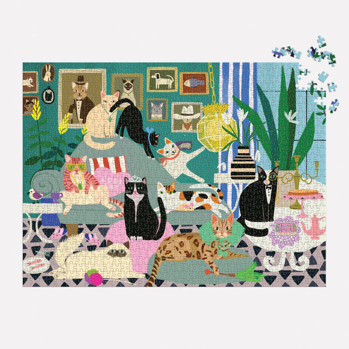 The Anne Bentley Glamour Cats 1000 Piece Puzzle features a vibrant room of stylish cats relaxing among plants, books, and art, with a few pieces missing in the upper right corner.
