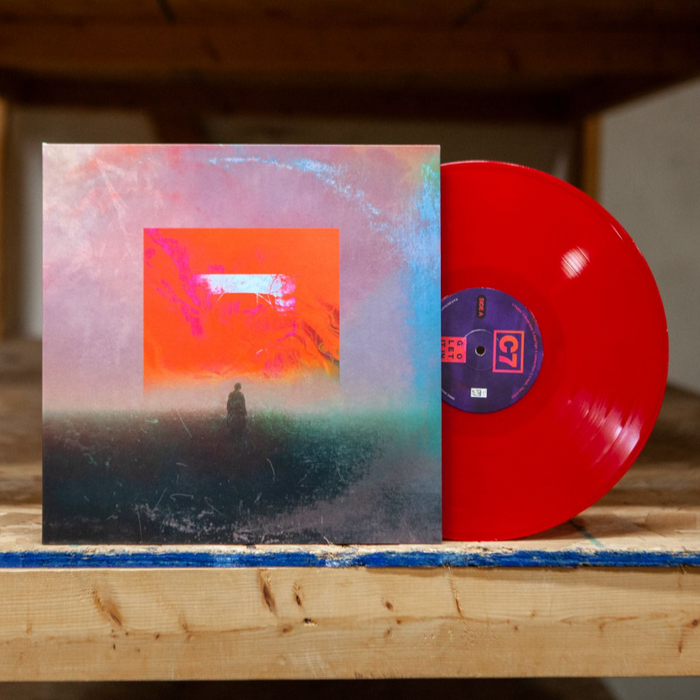 Vinyl is Red on side A. The jacket is a hazy image of a man standing underneath a red square.  The ground the man is standing on is black with a pink sky. The vinyl and jacket are propped against a wooden shelf.