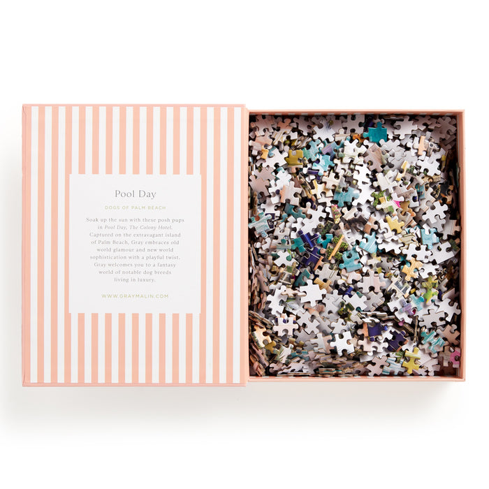 A striped pink and white box holds the Gray Malin Pool Day Palm Beach 1000 Piece Book Box Puzzle. The lid displays Gray Malin’s Pool Day design, mentions The Colony Hotel, and includes the website address.
