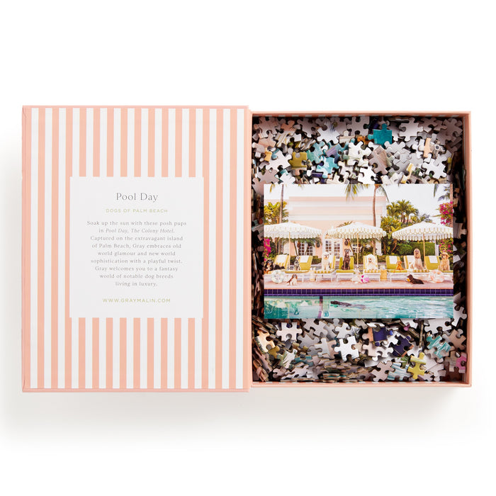 The Gray Malin Pool Day Palm Beach 1000 Piece Book Box Puzzle, by Gray Malin, comes in a striped pink and white box and features dogs relaxing poolside with umbrellas and palm trees in the signature Gray Malin style.