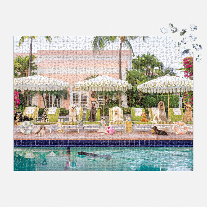 The Gray Malin Pool Day Palm Beach 1000 Piece Book Box Puzzle by Gray Malin shows dogs relaxing on poolside chairs under striped umbrellas at The Colony Hotel, with palm trees and a pink building; a few pieces are missing in the top right corner.