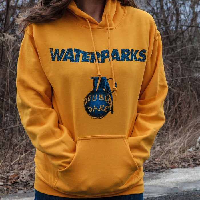 Yellow pullover hoodie with WATERPARKS written across the chest in dark blue lettering, Below that is a drawing of a dark blue grenade that says DOUBLE DARE in yellow writing on it. An individual is modeling the hoodie standing on the road with trees in the background.