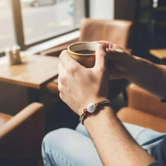 Handmade Tree of Life leather wrap bracelet worn by a person holding a coffee cup in a cozy cafe setting.