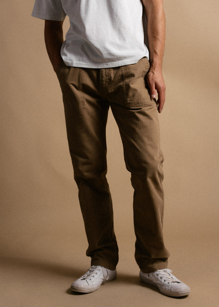 Man in a white t-shirt and brown pants stands casually against a beige background, wearing white sneakers.