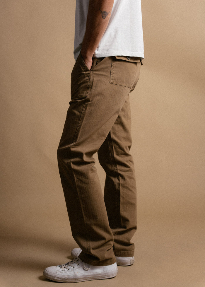Man in brown pants and white sneakers stands sideways, hands in pockets, against a beige background, showcasing casual style.