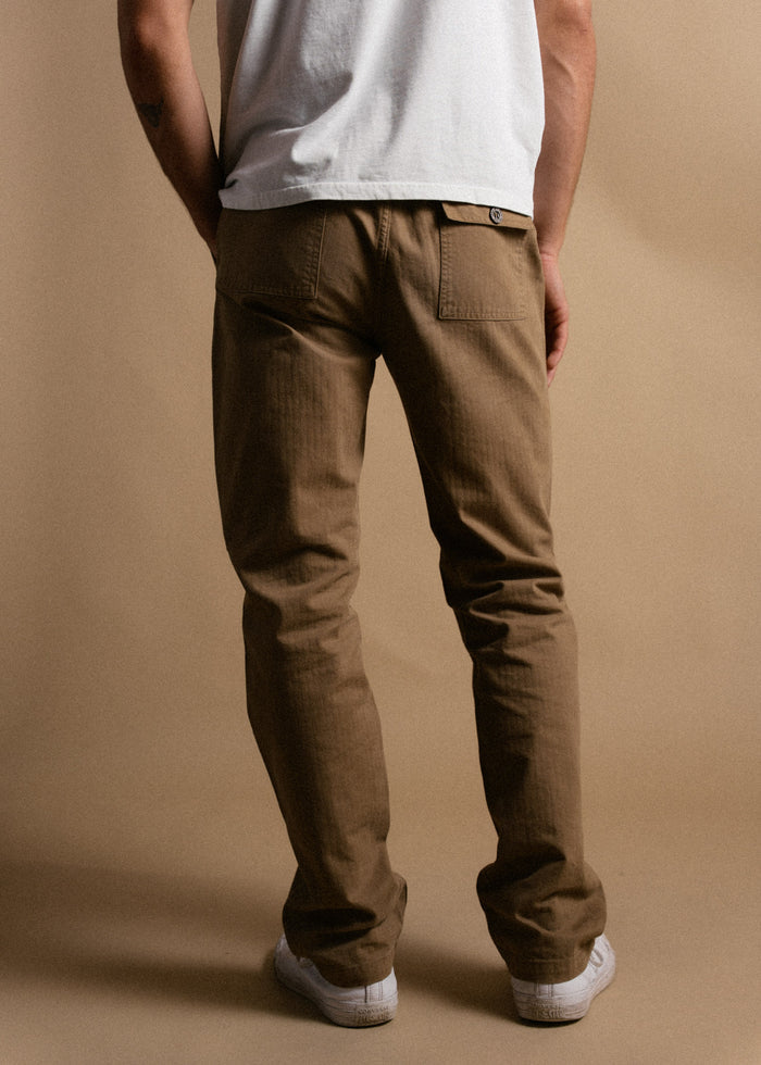 Man in white shirt and brown pants stands against a beige background, hands by sides, wearing white sneakers.