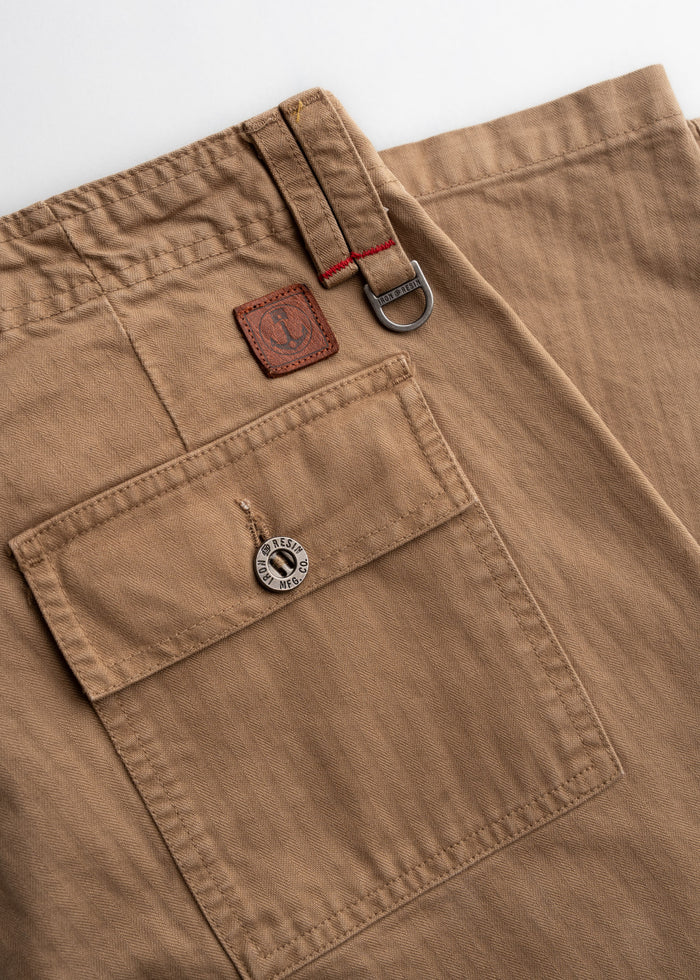 Close-up of khaki pants with a buttoned pocket, leather patch, and metal ring detail. Red stitching adds contrast to the design.