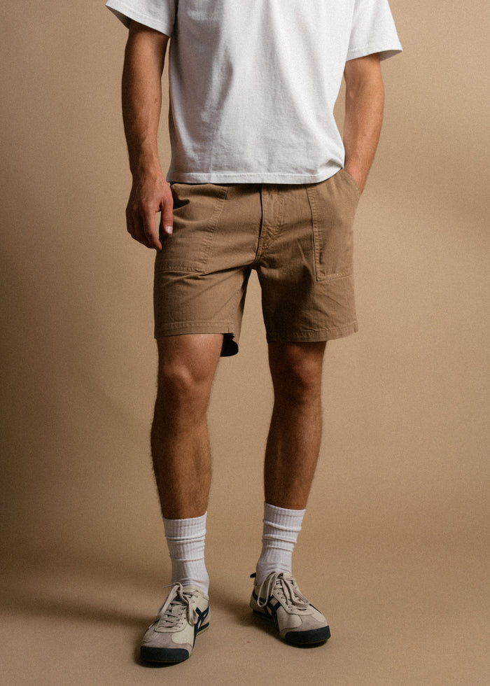 Man in white shirt, brown shorts, white socks, and sneakers stands against a beige background, hands in pockets, casual style.